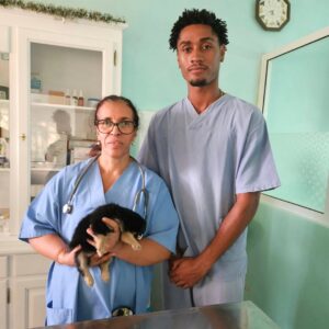 A female vet in blue scrubs with a stethoscope holding a small black-and-white puppy in a clinic, standing beside a male colleague in light blue scrubs.